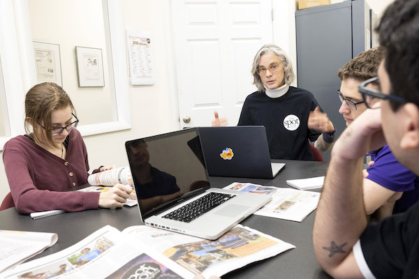 A group of students sit at a table with laptops discussing journalism and articles with a professor wearing a blue sweater.