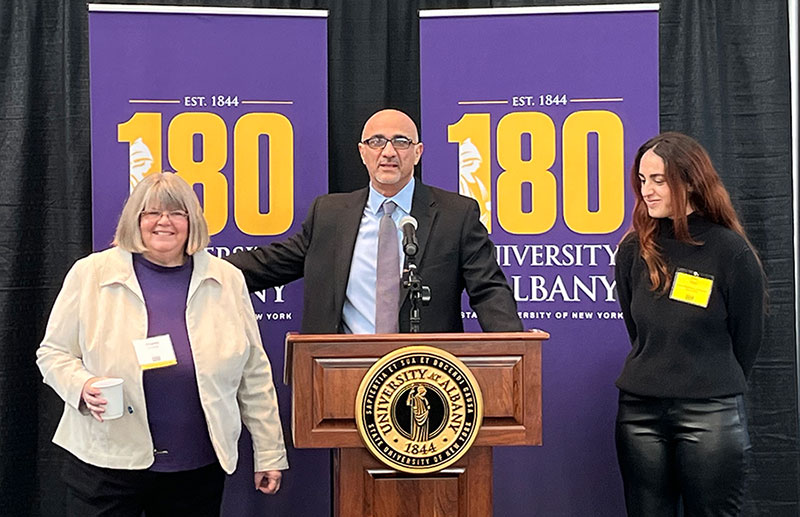 Virginia Goatley, Fardin Sanai and a UAlbany student stand at a podium in front of 180th Anniversary Banners.