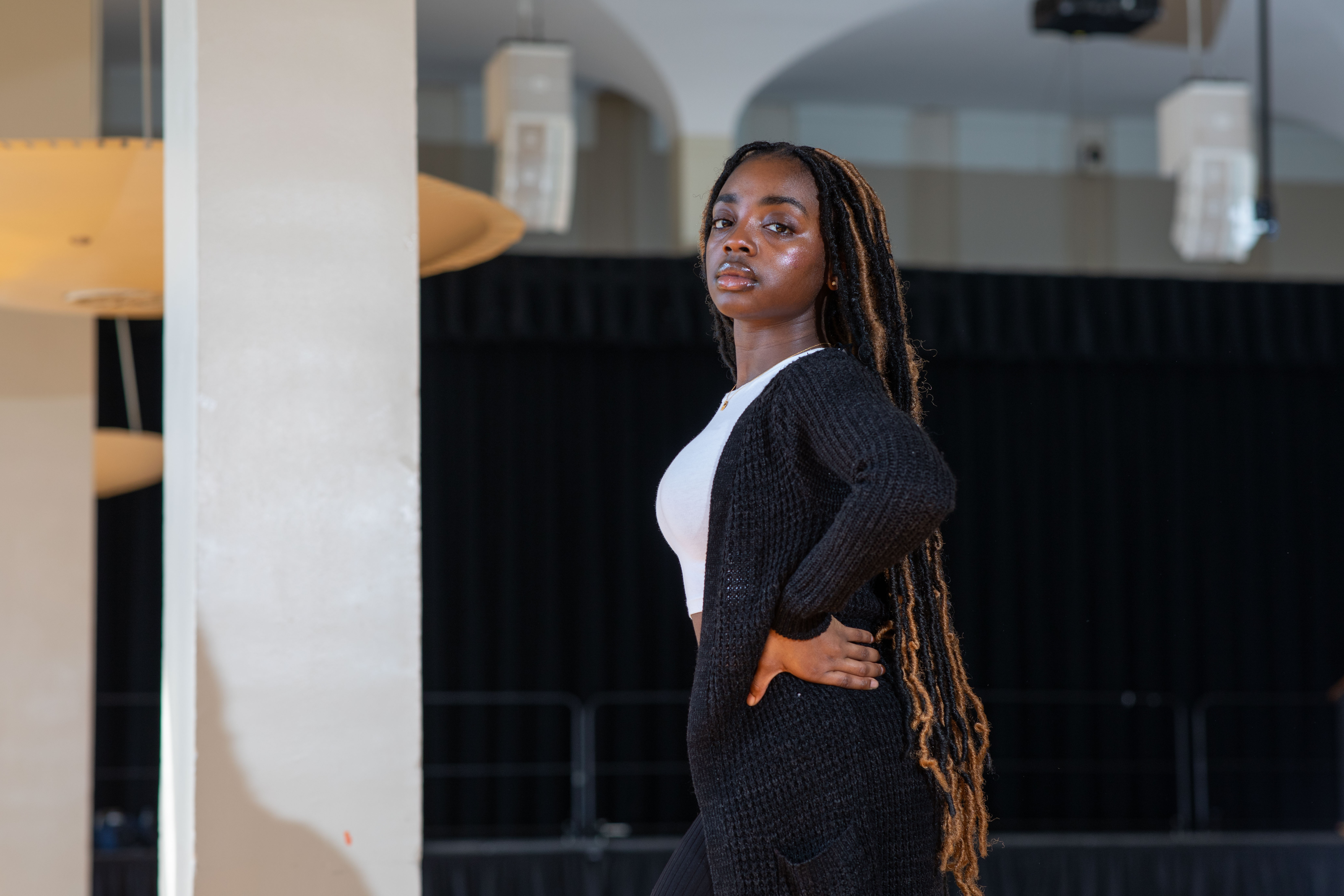 A woman with long black and brown hair in braids wears a white shirt and black cardigan and poses with a hand on hip next to a white column