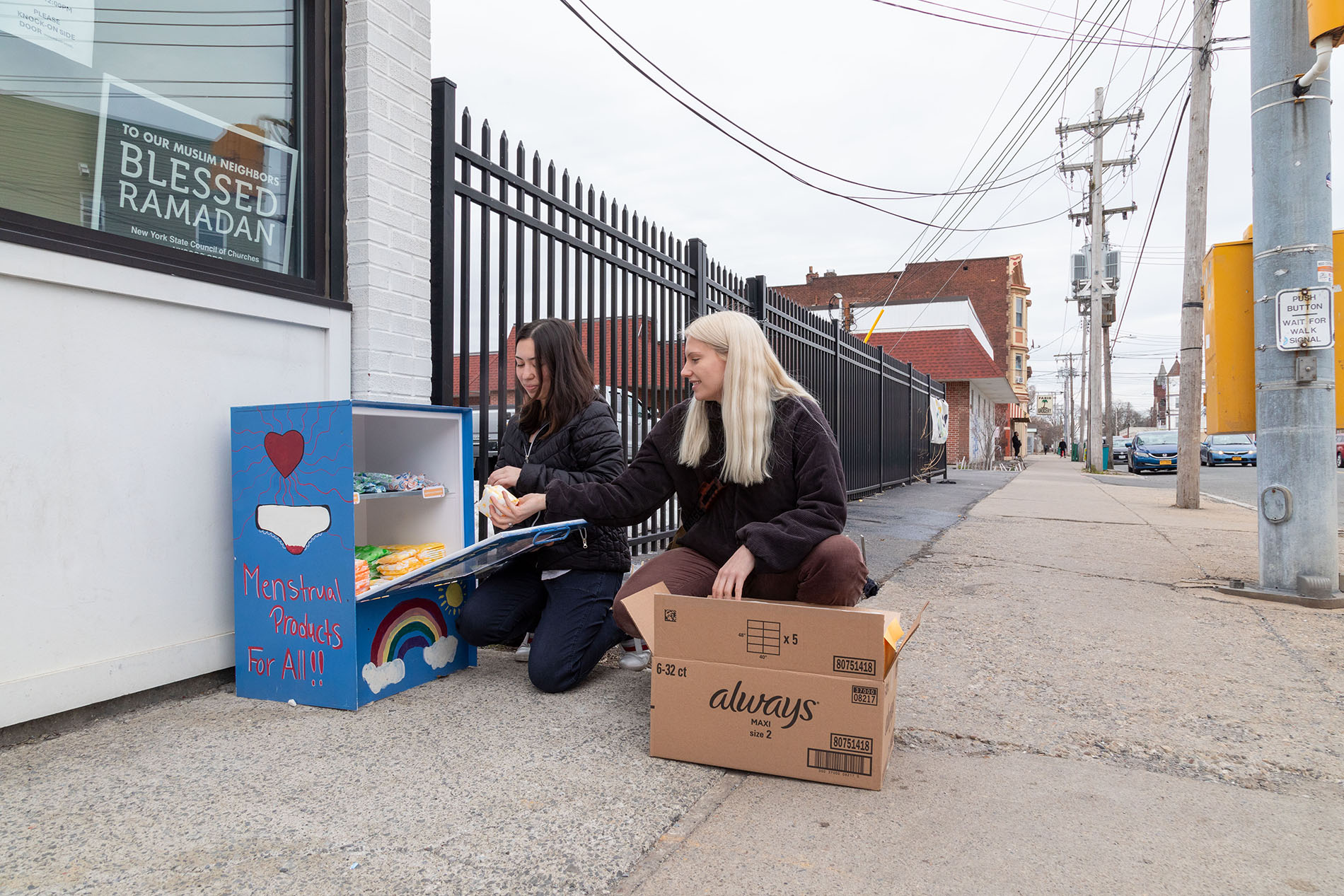 Two young women crouch on a sidewalk next to a blue metal box which they are stocking with a variety of period products.