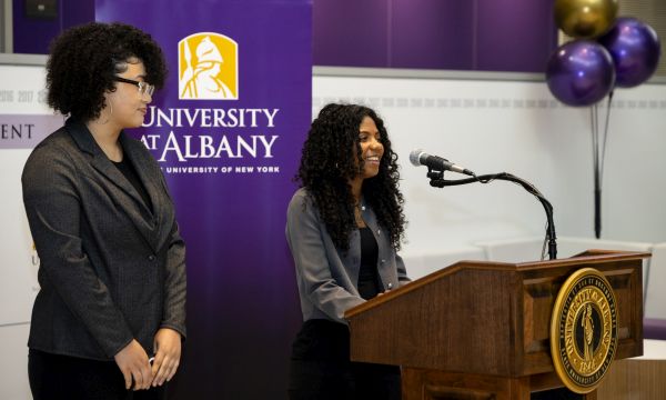 two women stand near a podium in front of a banner reading University at Albany