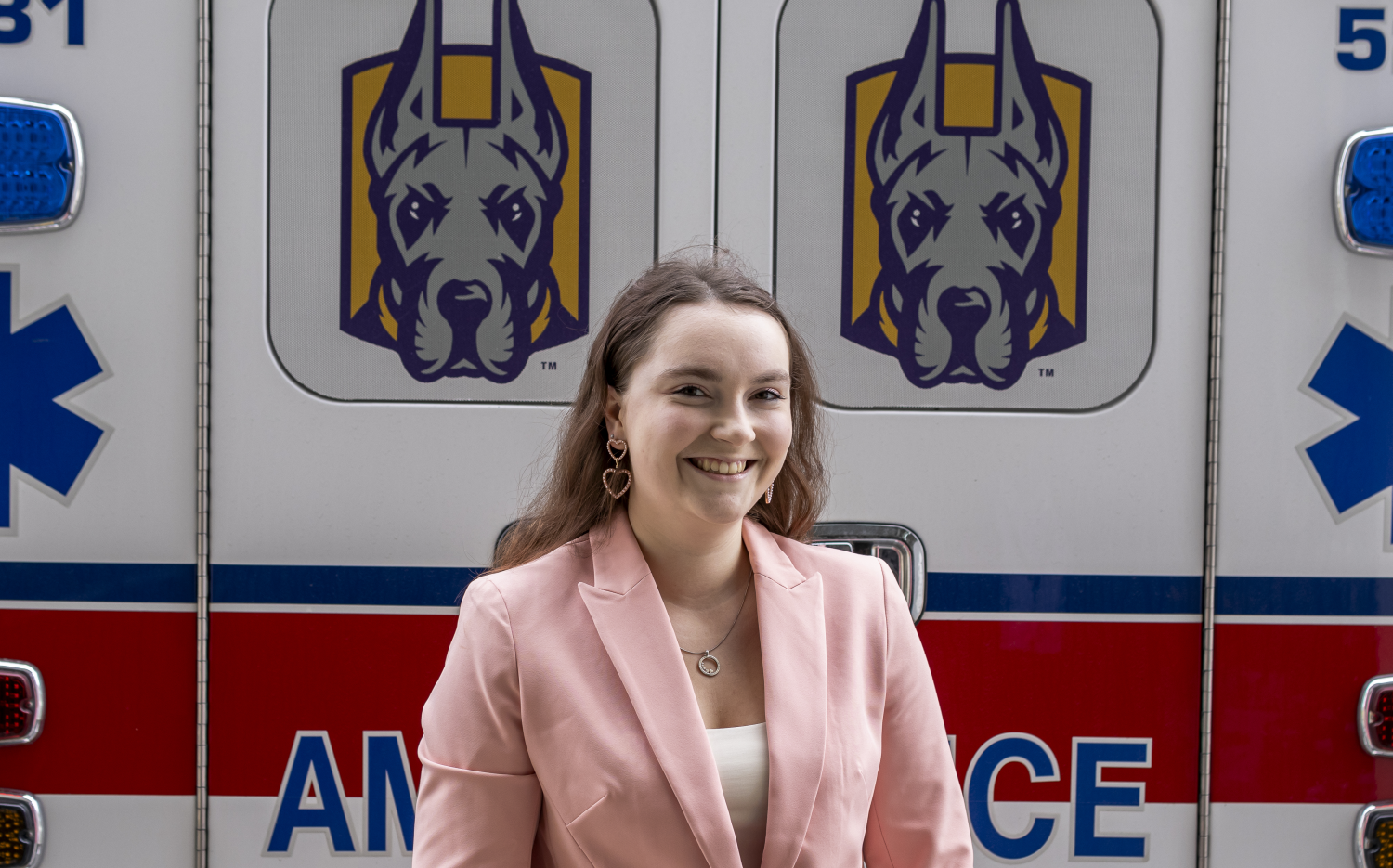 A smiling woman in a pink jacket stands in front of an ambulance that has pictures of UAlbany's mascot on it
