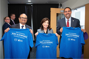 From left to right (Kesha Kesavadas, Gov. Kathy Hochul and SUNY Chancellor John B. King Jr. stand smiling should to shoulder holding up blue t-shirts for the inaugural SUNY AI Sympisium.