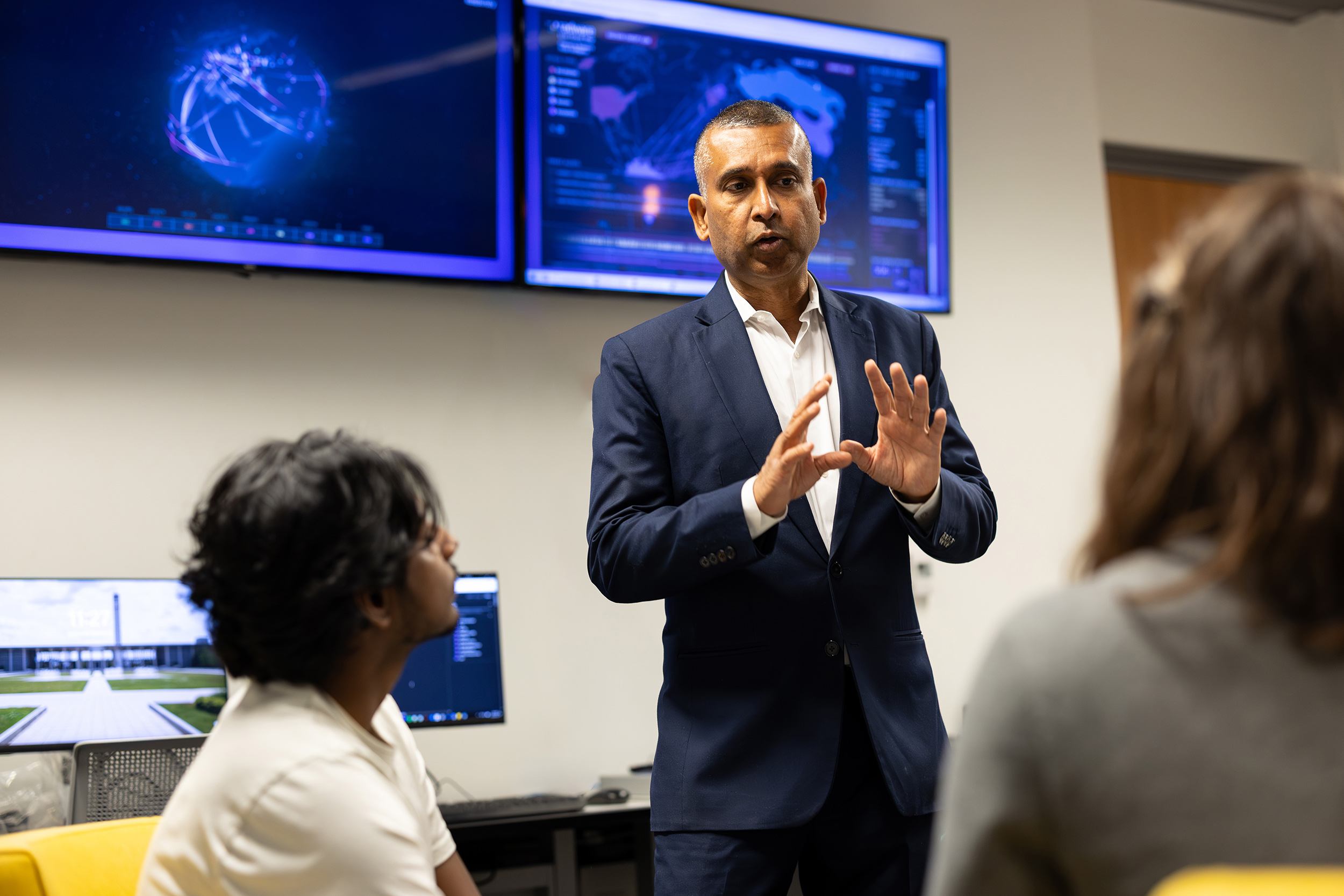 A professor with a blue blazer stands before a small group of students with monitors displaying a digital map of the world in the background indoors.