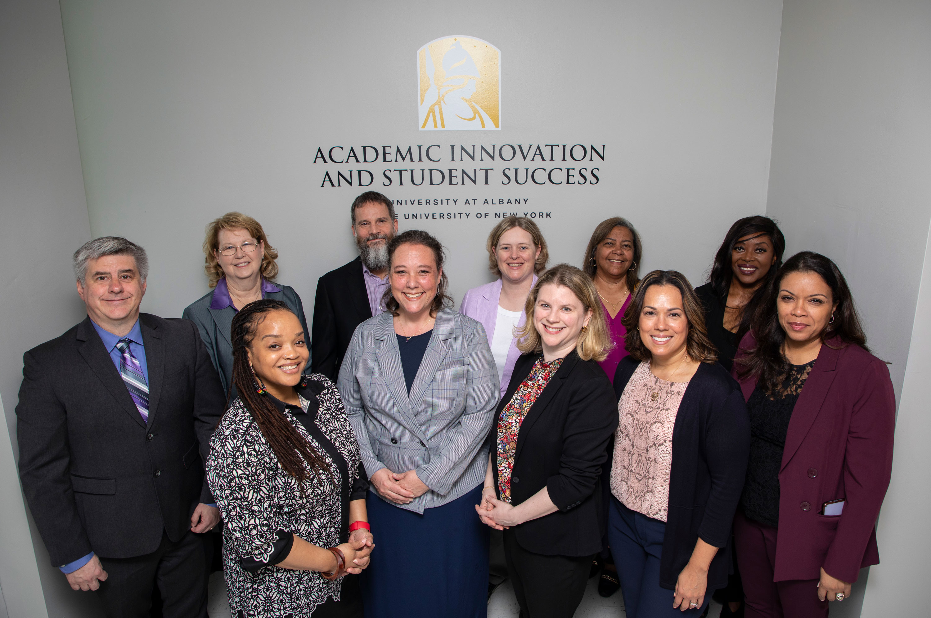 Eleven of the AISS leaders pose for a photo in front of a wall with a sign with the UAlbany logo and the words, "Academic Innovation and Student Success, University at Albany, State University of New York."