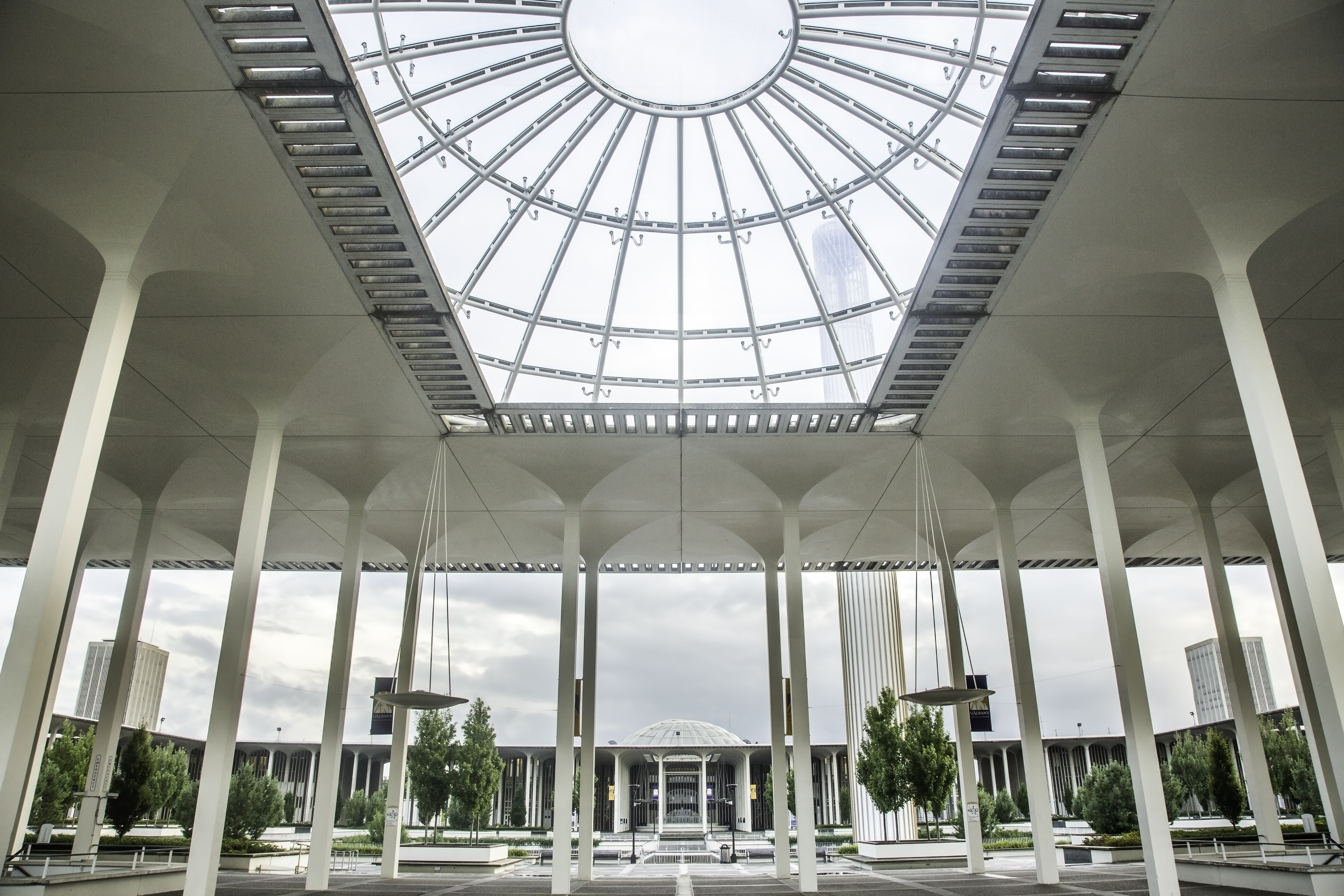 UAlbany's main campus shot with tall columns holding up a windowed roofing structure. The campus fountain can be seen in the background. It is a cloudy but beautiful day on campus.