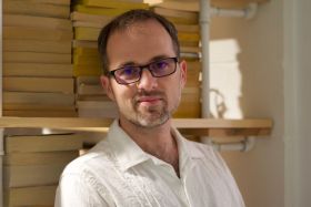 A bespectacled man with a goatee leans against a stack of books.