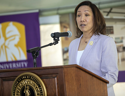 UAlbany Provost Carol H. Kim speaking at a podium inside of a tent with a UAlbany flag in the background.