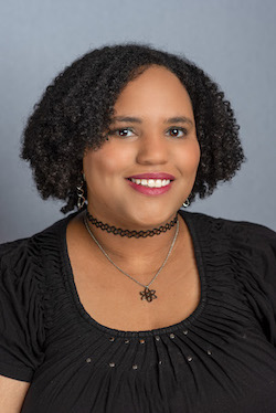 A young woman with short curly black hair wears a black top, black choker and a necklace and smiles for a portrait against a gray backdrop.