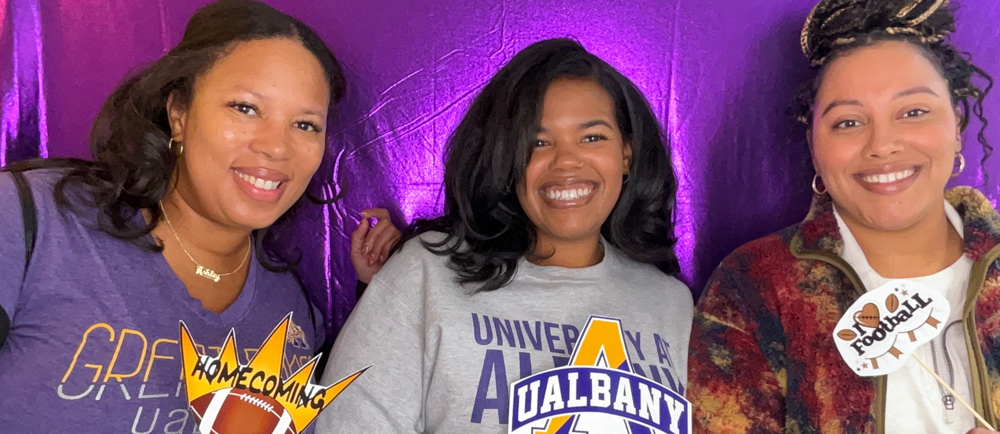 Three UAlbany alumni wearing purple and gold pose for a picture inside a photo booth.