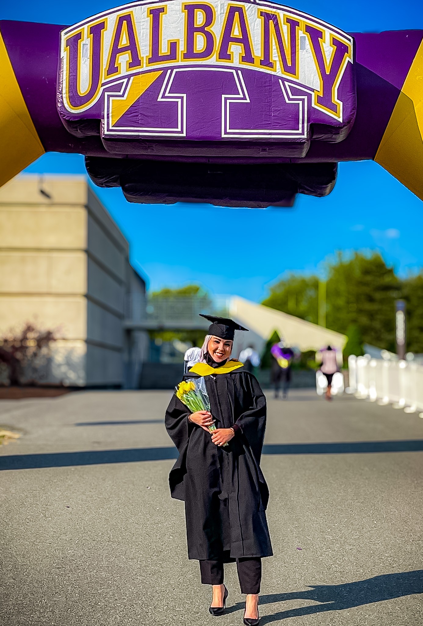 Amaal Alharbi smiles and poses for a photo under a blow up UAlbany arch. She is wearing a graduation cap, gown and graduate cowl, and holding a bouquet of flowers.