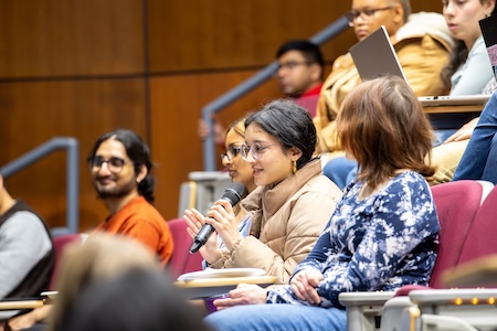 A young woman seated in an auditorium surrounded by other people speaks into a microphone
