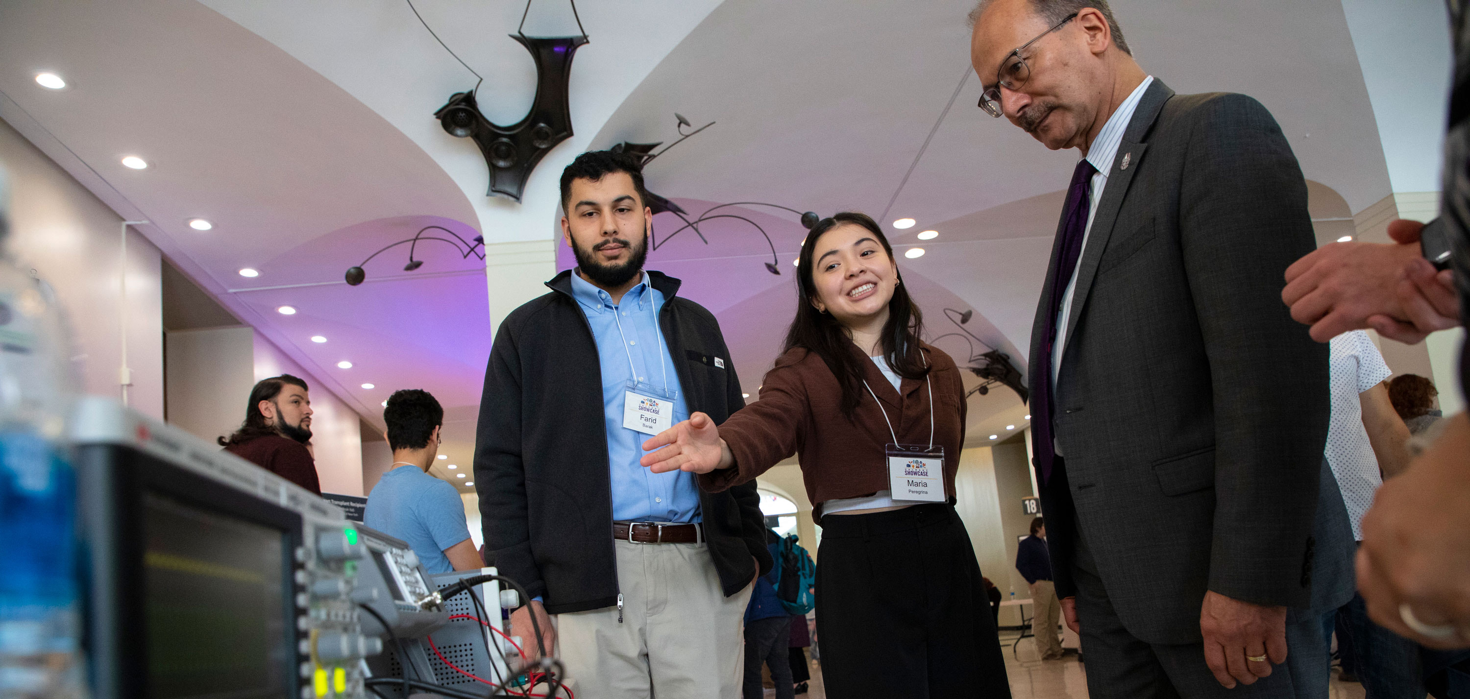 Two students stand in front of a table with audio equipment. One student gestures toward the table while speaking to UAlbany President Havidán Rodríguez.