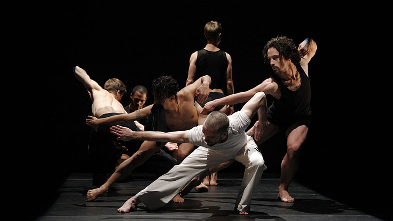 many dancers in a clump lunge to one side while a single dancer stands alone in background