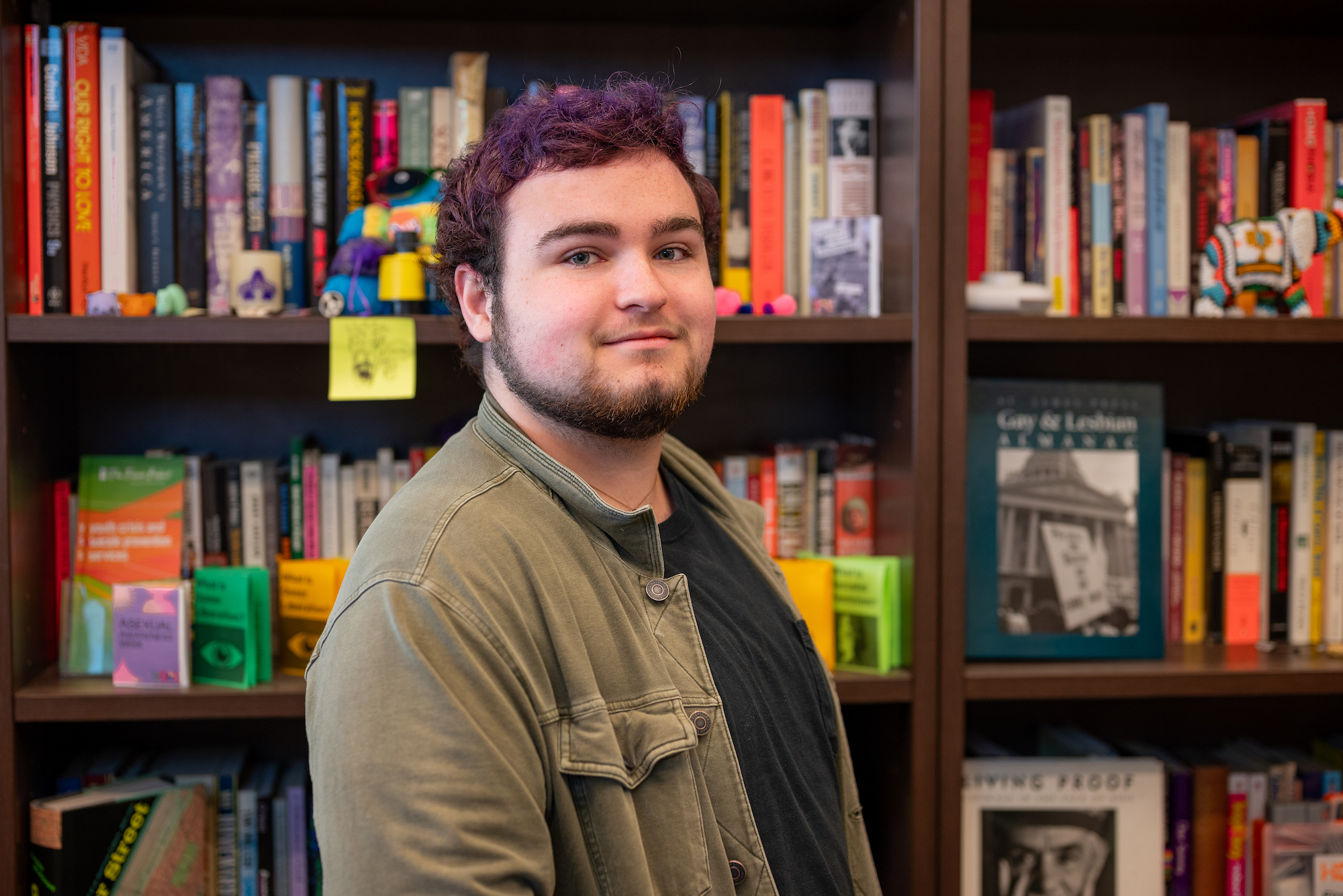 A person with short purple hair and a goatee smiles for a portrait in front of bookshelves.