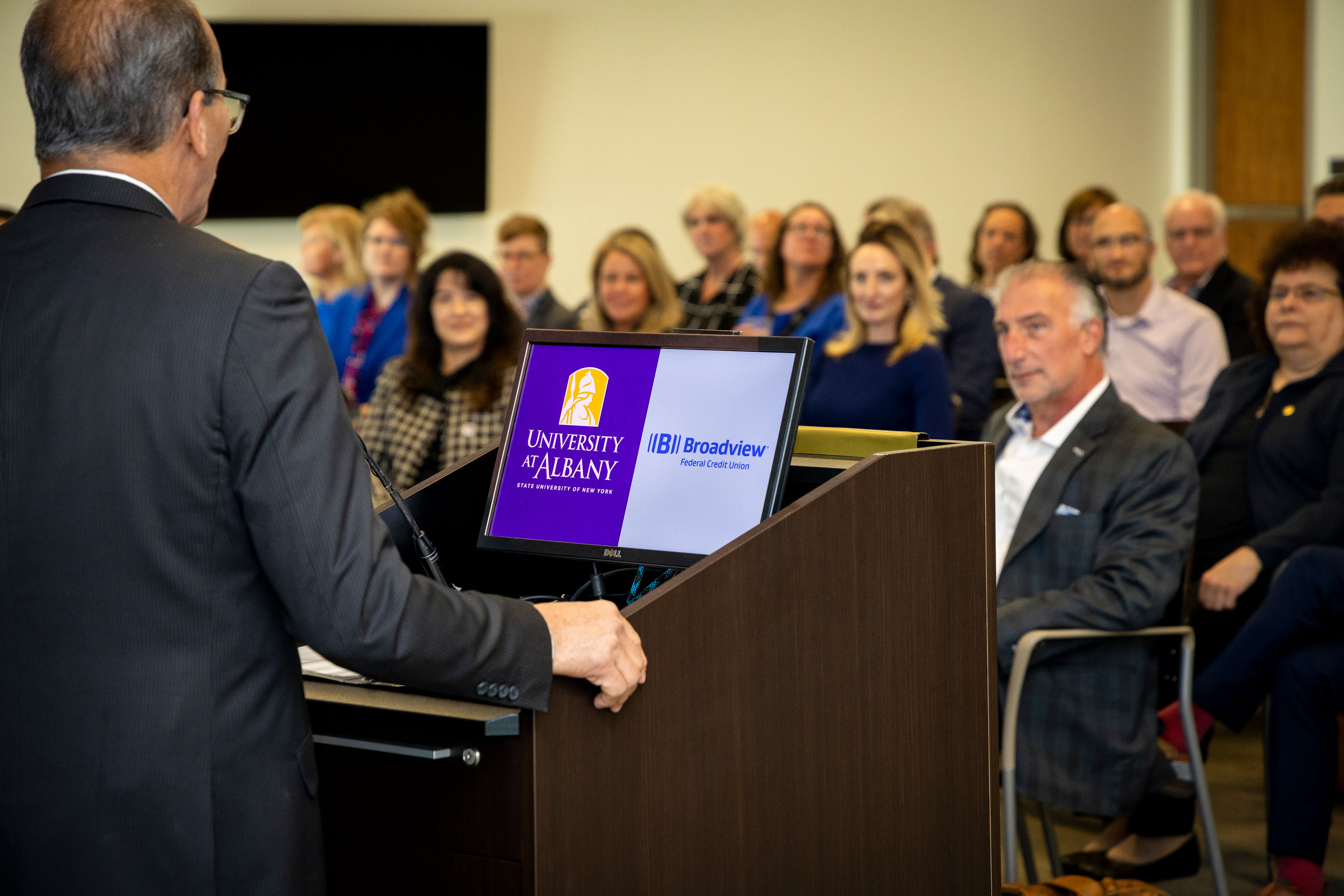 UAlbany President Havidán Rodríguez stands at a podium with computer displaying the UAlbany and Broadview logos, as he speaks to a gathered group during the partnership signing ceremony.