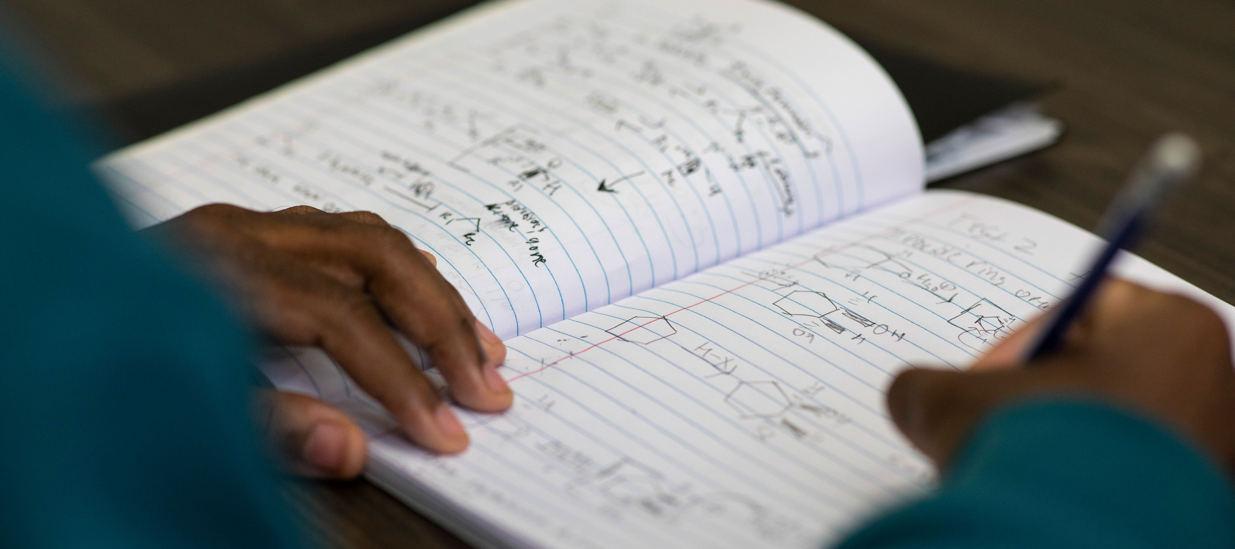 A student writes in a notebook that contains STEM notes and equations.