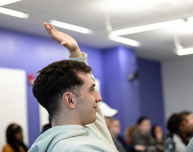 A student in a light-colored hoodie raises his hand in a crowded classroom.