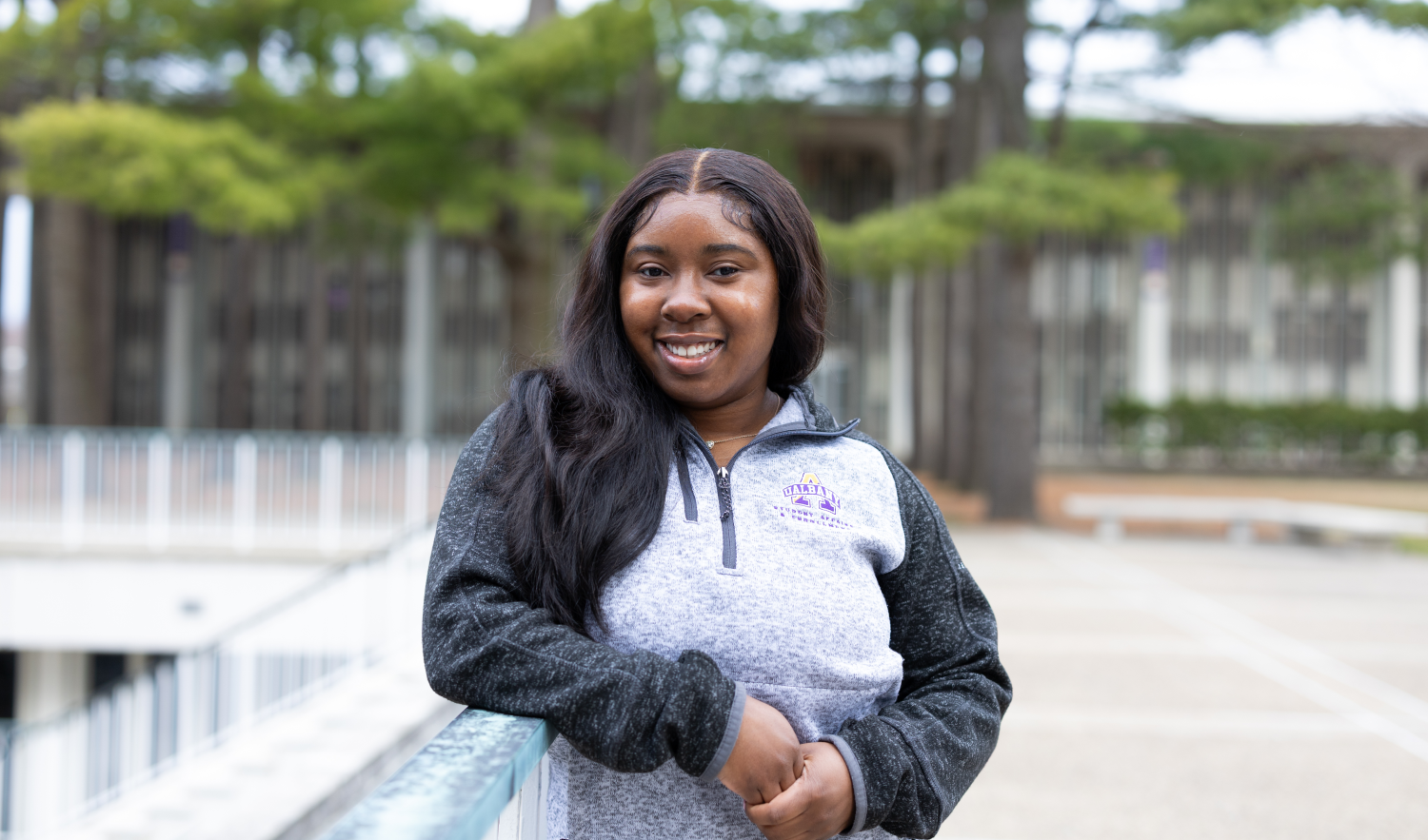 a woman in a sweatshirt stands outside, in front of a row of trees blurred in the background