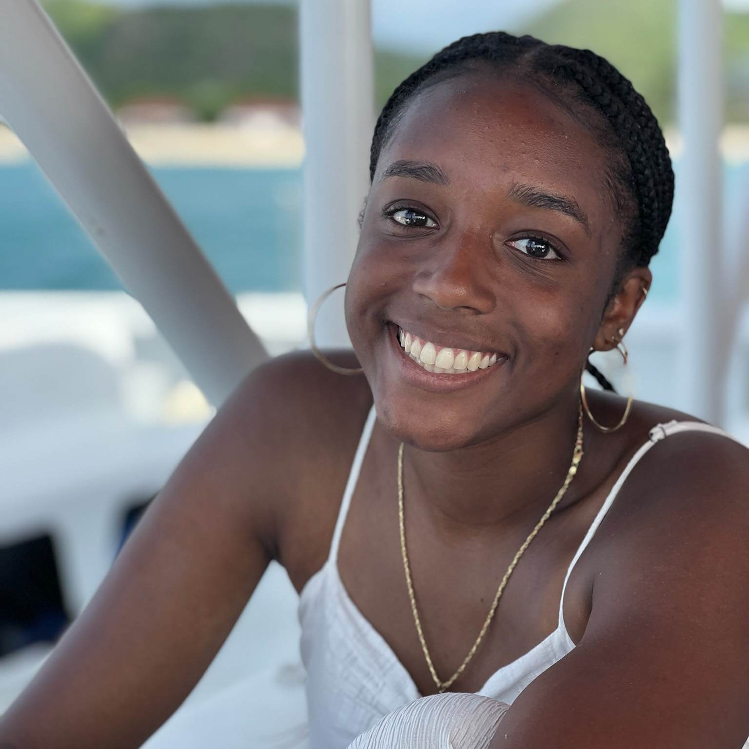 Outdoor portrait of Camryn Beckles, smiling and seated. Beckles is wearing a white top, gold hoop earrings and gold necklace.