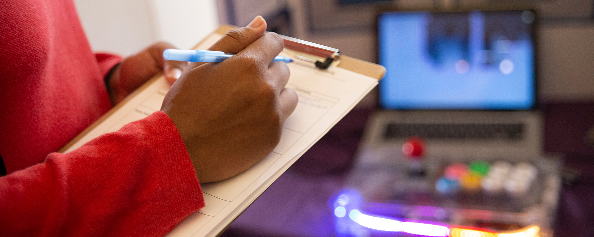 A hand poised to write on a clipboard, with a computer system visible in the background