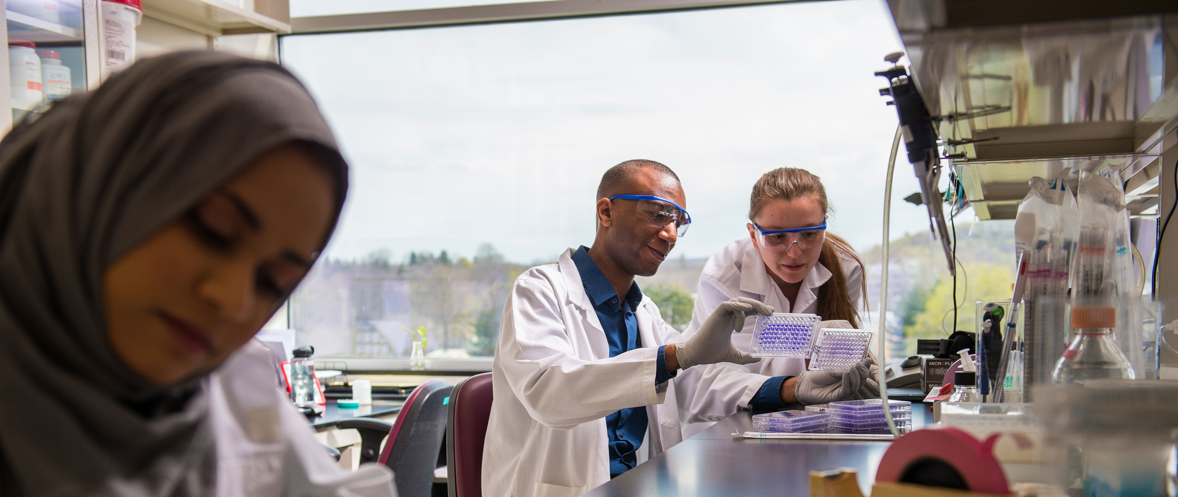 Two researchers in white lab coats and protective glasses examine a specimen in the background, while a third researcher writes in a notebook in the foreground.