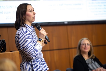 A woman in a blue and white striped button down speaks into a microphone on a stage as another woman watches