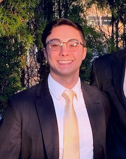 A young person wears glasses, a dark suit and gold tie and smiles for a photo outdoors at night.