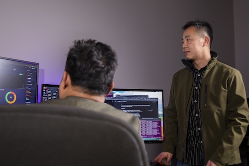Two men in a computer lab sit and stand near a bank of computers.