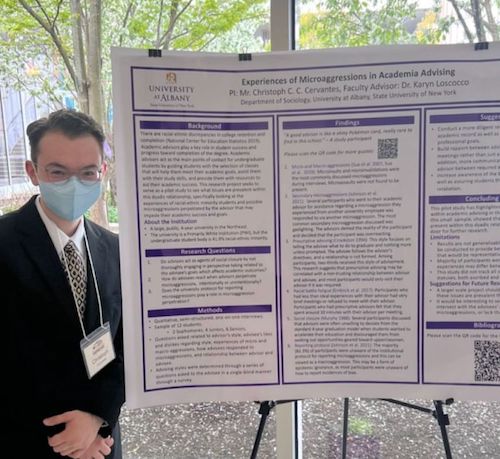 A young man with short brown hair, glasses and a face mask poses next to a research poster.