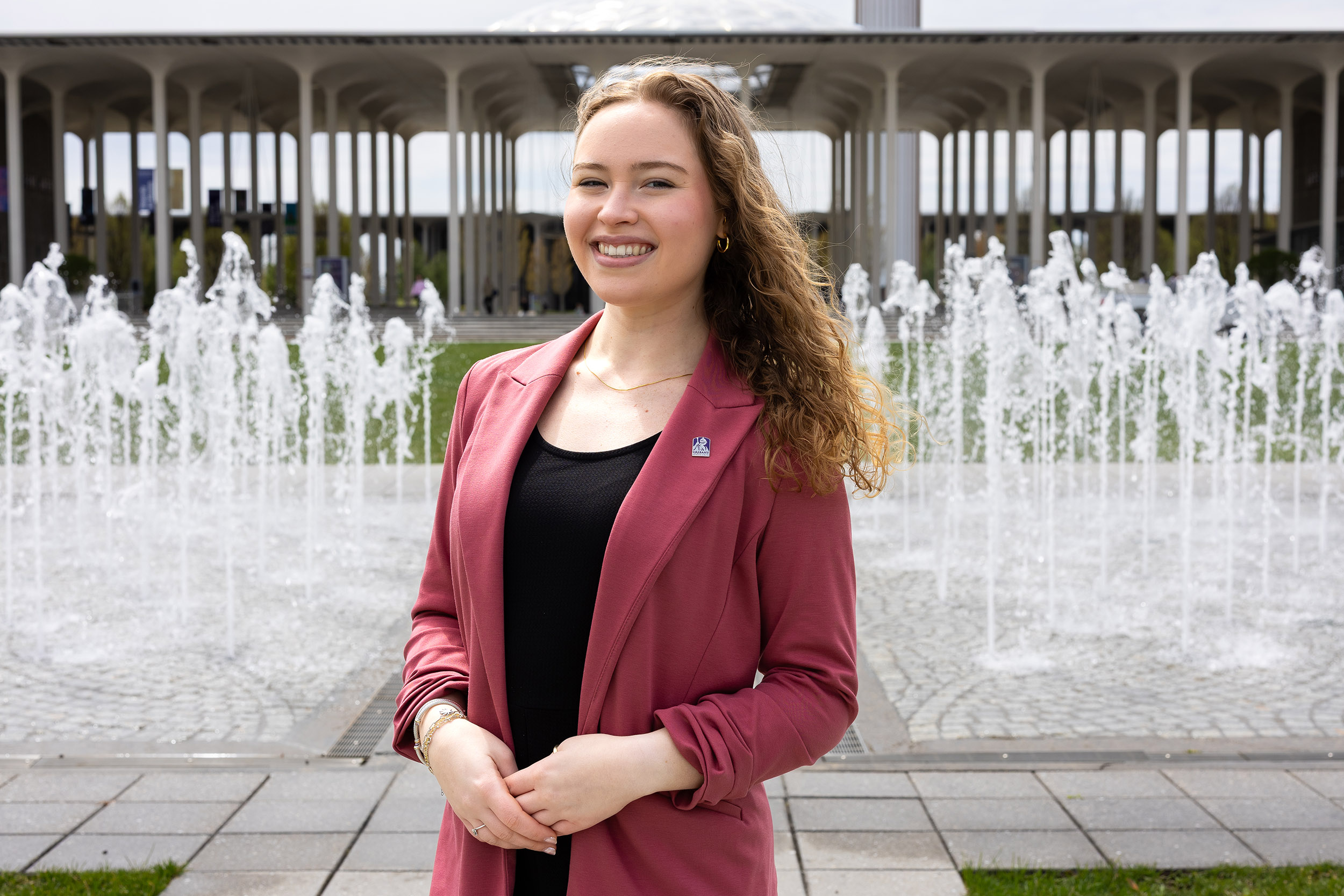A portrait of UAlbany student Sophie Coker as she stands in front of the entry plaza fountain