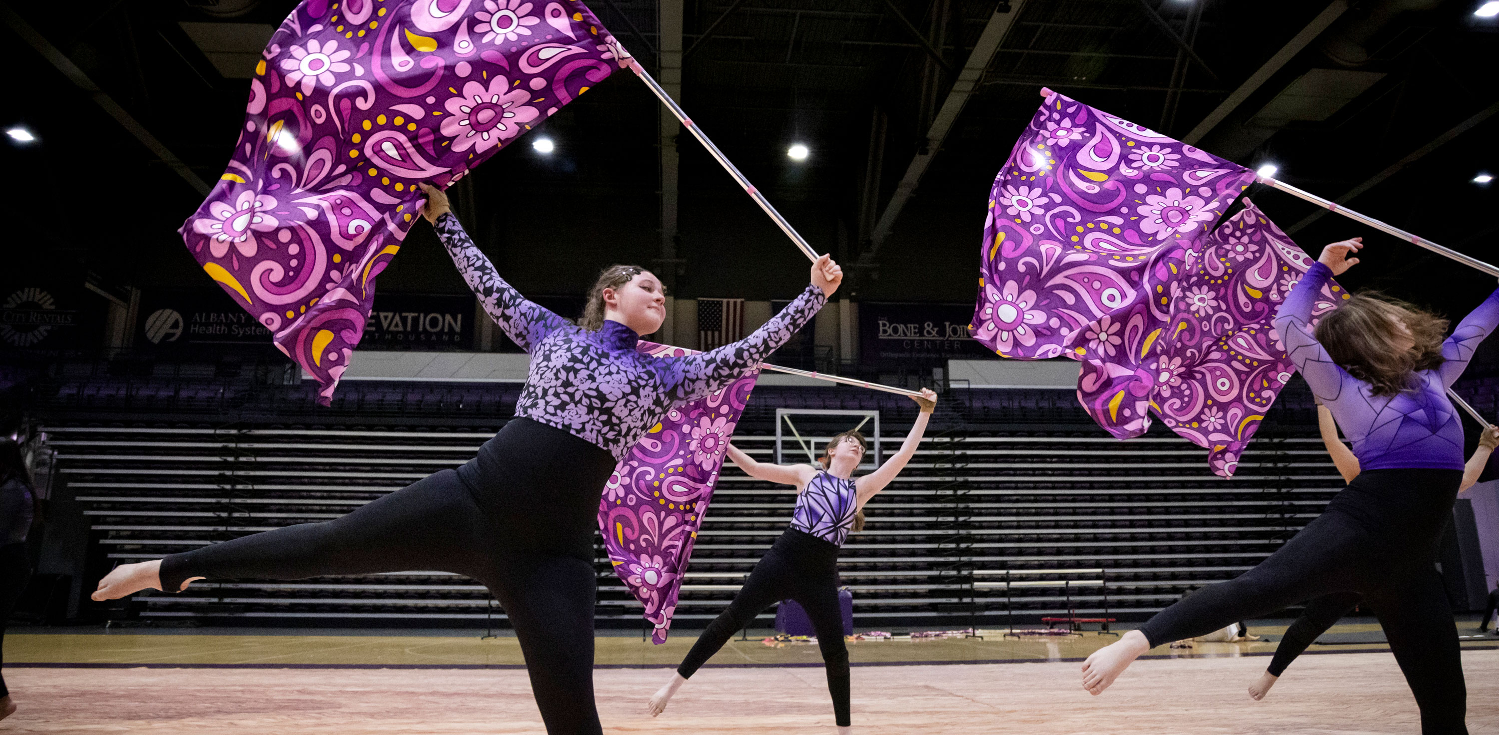 Four members of the UAlbany Winter Guard balance on one foot in matching outfits as they wave purple flags inside an arena.