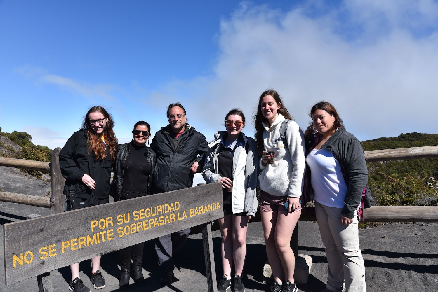 A small group of students and a faculty member stand beside a sign in Spanish. They are outdoors and a clear blue sky is behind them.