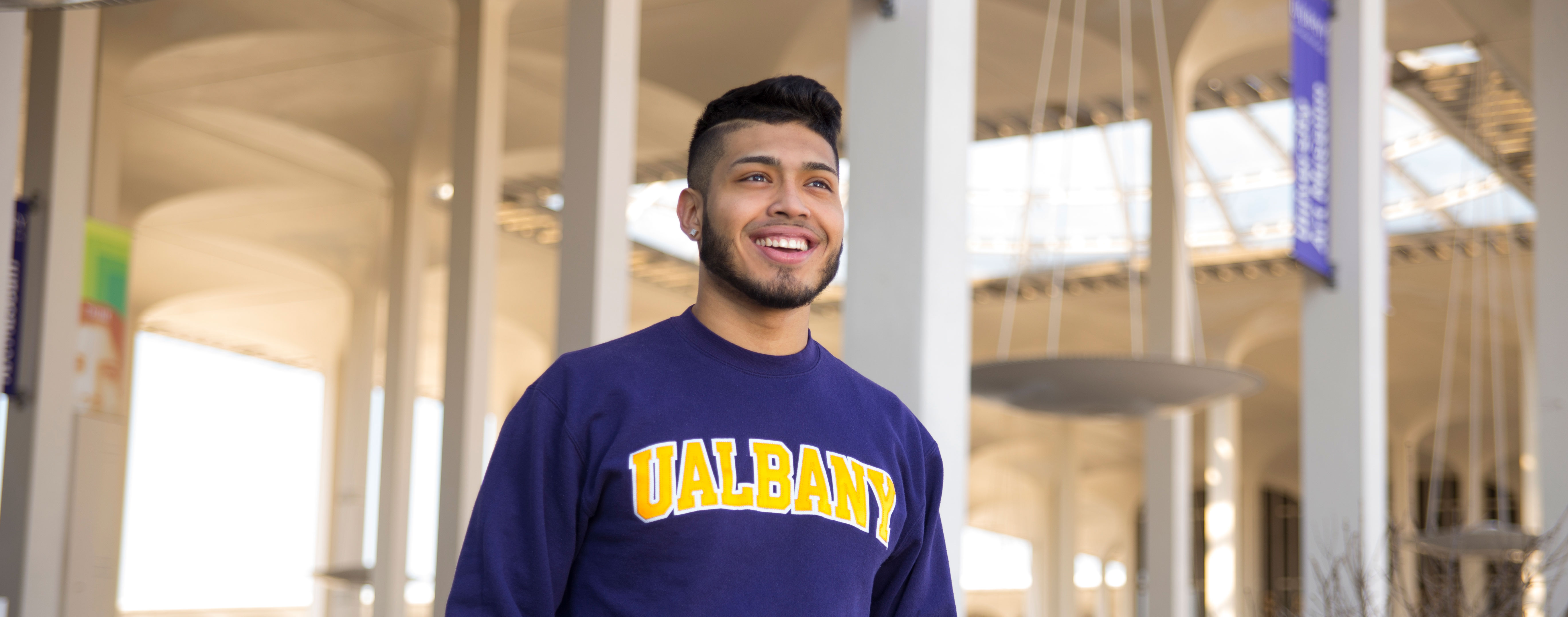 A student wearing a purple UAlbany sweatshirt smiles and poses for a photo on campus, with the Podium arches visible behind him
