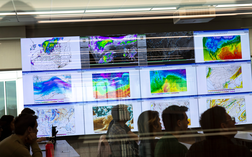 A classroom full of students is seen through a window, while the wall behind them is made up  of screens displaying a variety of weather maps and data.