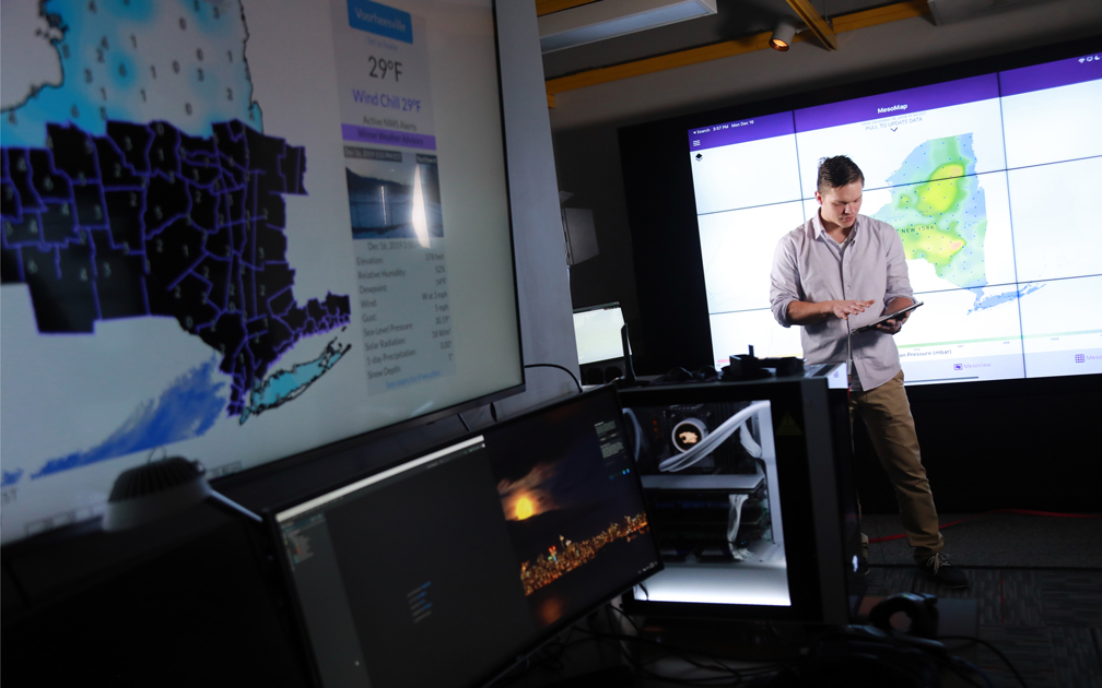 A grad student in a button up shirt and khakis stands in front of a wall-sized monitor displaying a weather map at UAlbany's xCITE Lab. He is surrounded by weather equipment, including a screen in the foreground that reads "29°"
