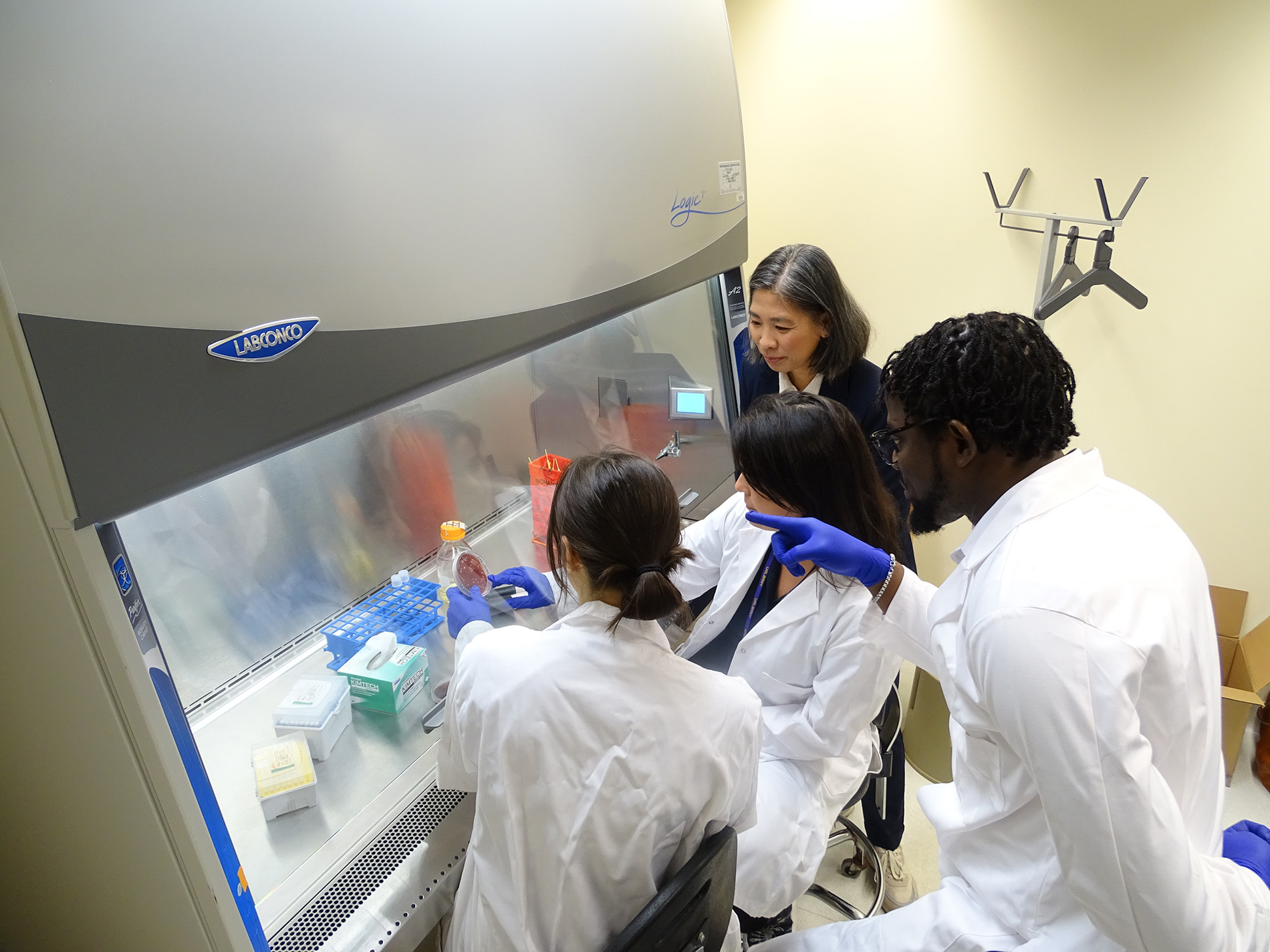 Three people wearing white lab coats (two women and one man) gather around a lab bench protected by a glass hood. A women wearing a navy blazer looks on. The lab bench contains test tubes, petri dishes and other plastic containers for biology research.