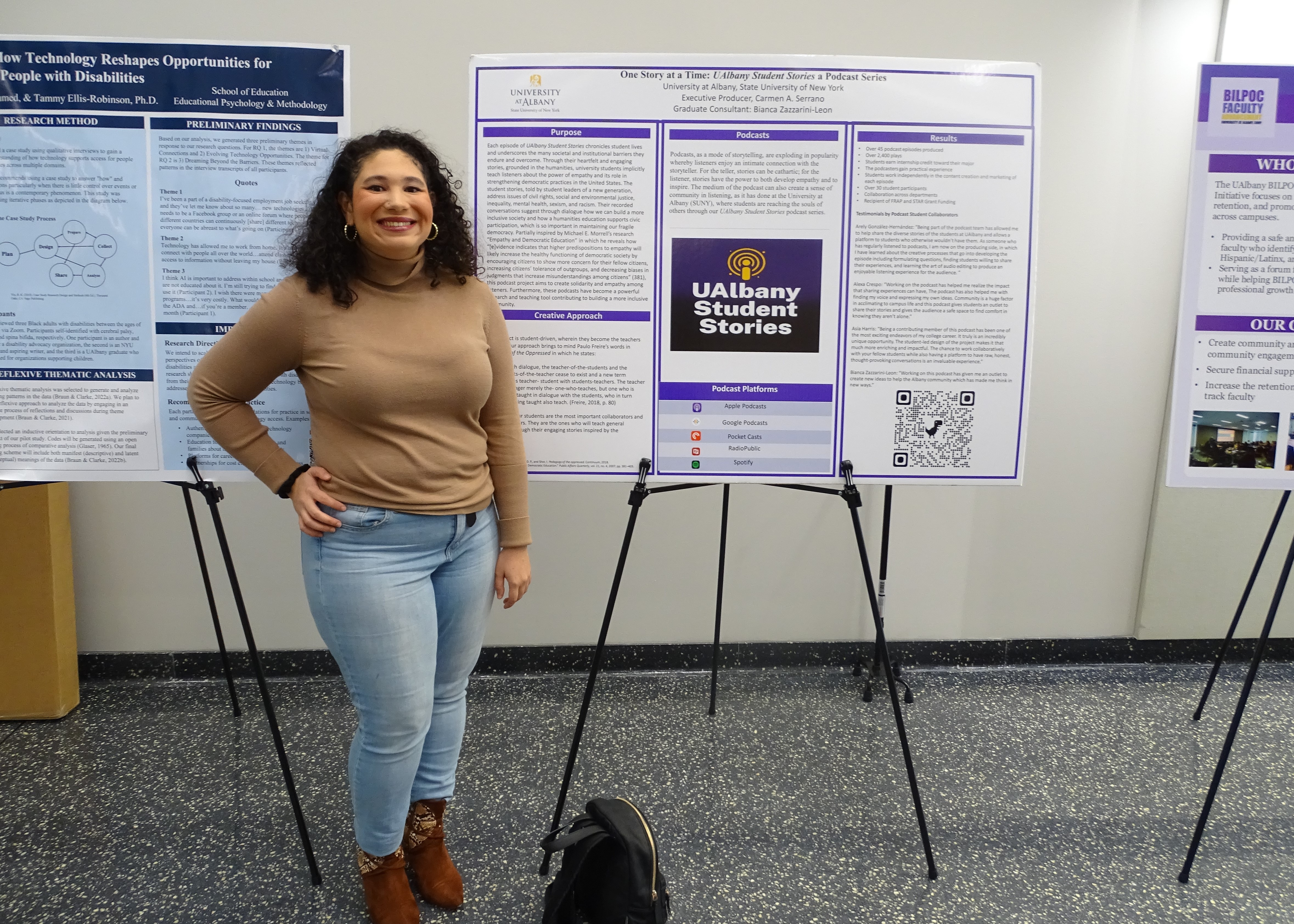A young woman with curly brown hair wearing a tan sweater and brown boots smiles in front of a poster about the UAlbany Student Stories Podcast.