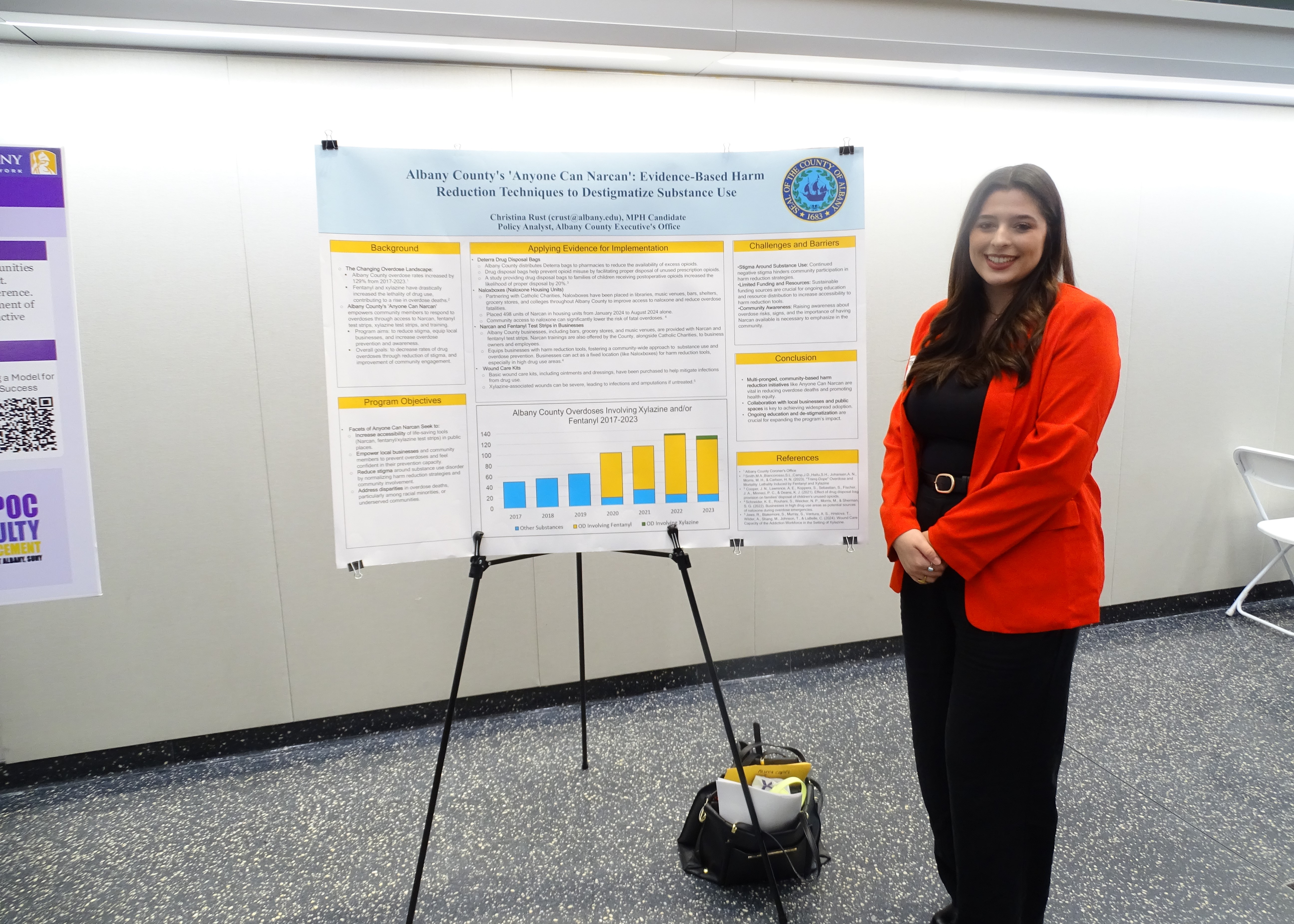 A young woman with long dark hair, wearing a red blazer and black pants smiles beside her poster titled "Albany County's 'Anyone Can Narcan': Evidence based harm reduction techniques to destigmatize substance use." 