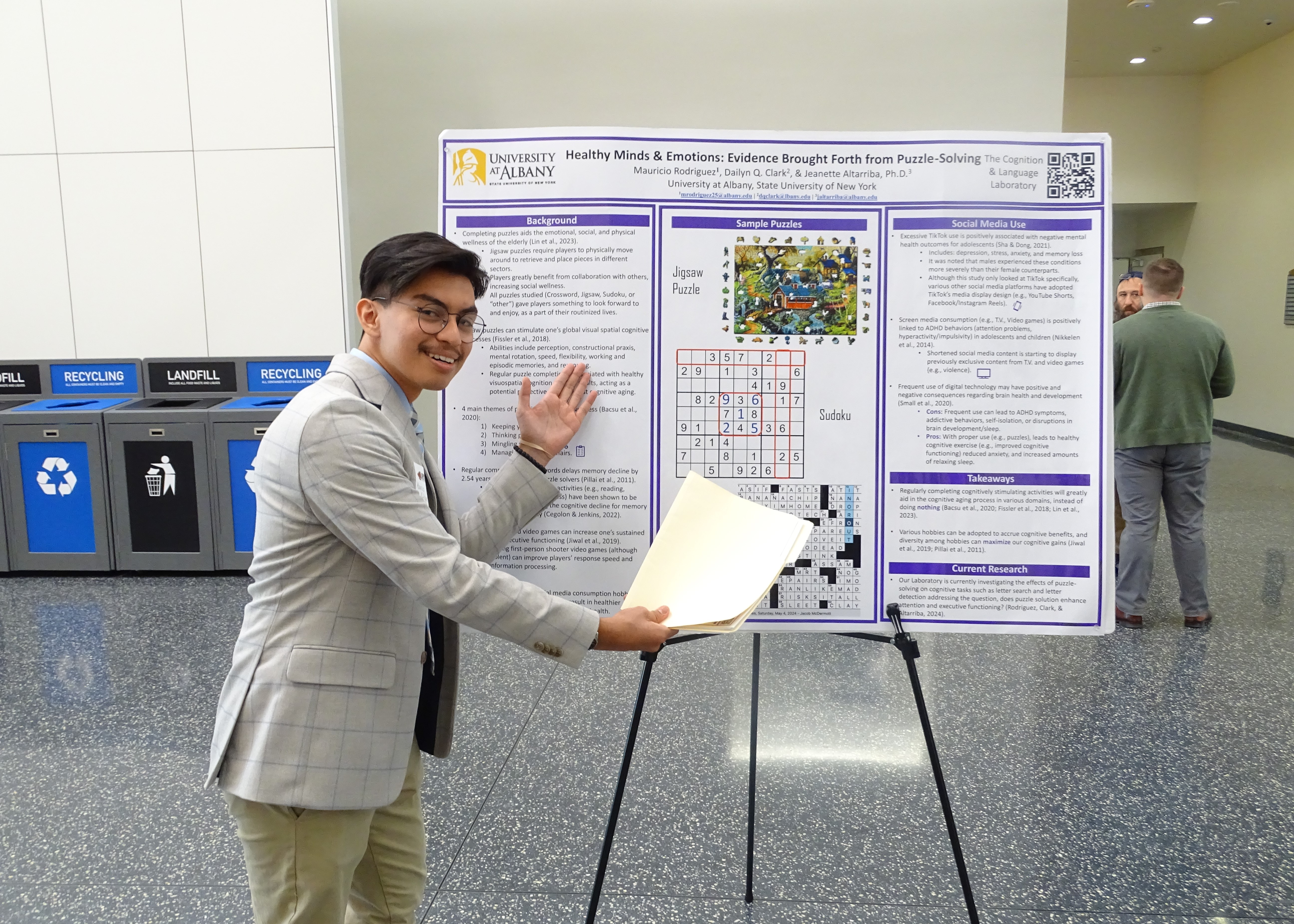 A young man wearing glasses and a grey blazer displays his poster titled "Healthy Minds and Emotions: Evidence Brought Forth from Puzzle Solving."