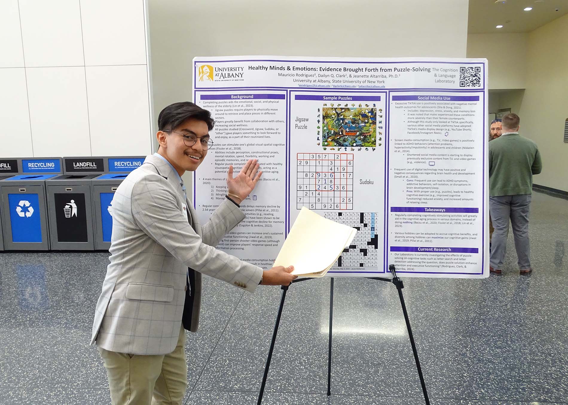 A young man wearing glasses and a grey blazer displays his poster titled "Healthy Minds and Emotions: Evidence Brought Forth from Puzzle Solving."
