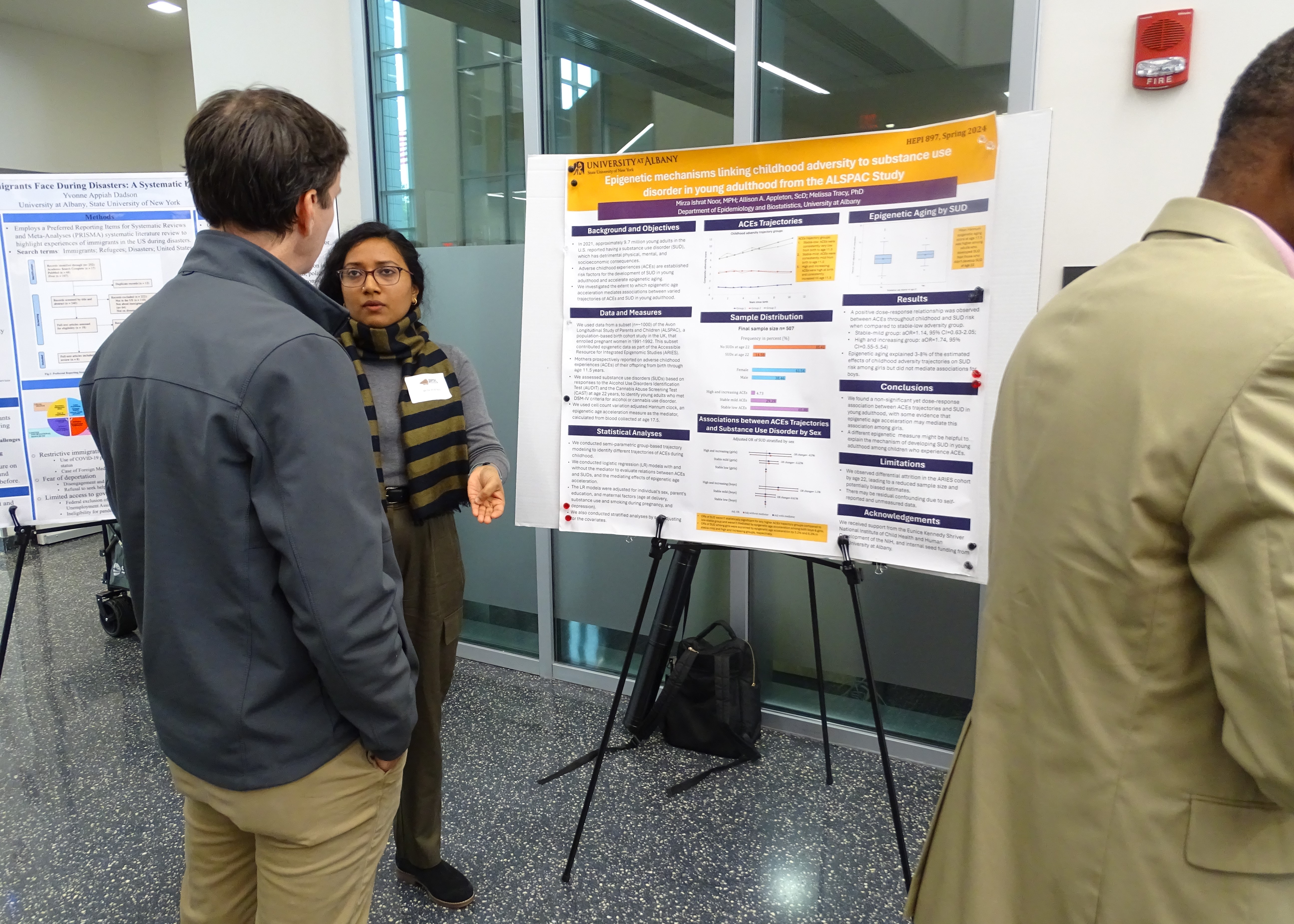 A woman in a striped scarf and glasses stands in front of a poster, speaking to a man in a grey jacket. Her poster reads: “Epigenetic mechanisms linking childhood adversity to substance use disorder in young adulthood from the ALSPAC Study."