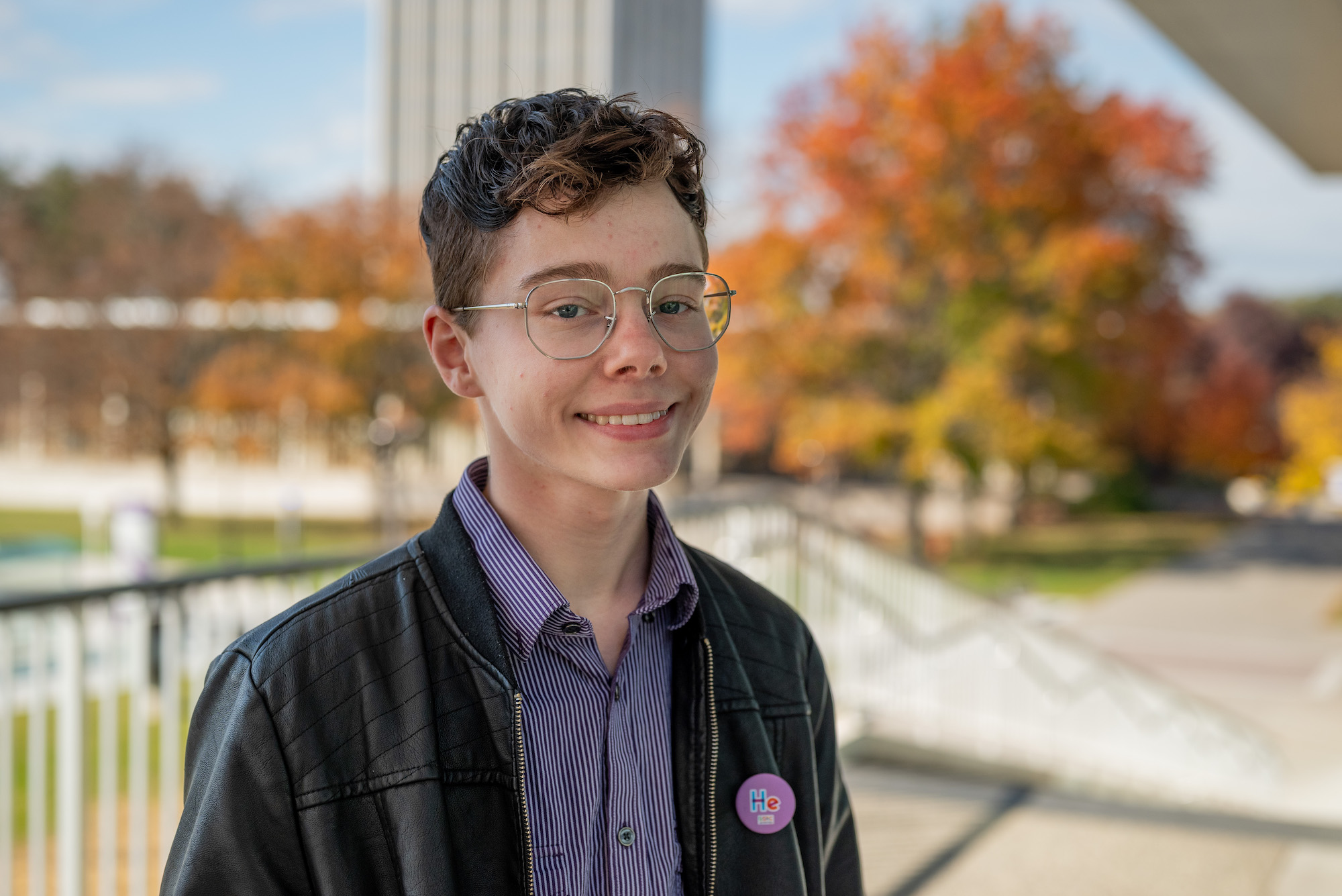 A young person with short curly hair and glasses wears a leather jacket and striped shirt and smiles for a portrait on the UAlbany campus. Fall foliage can be seen in the background.