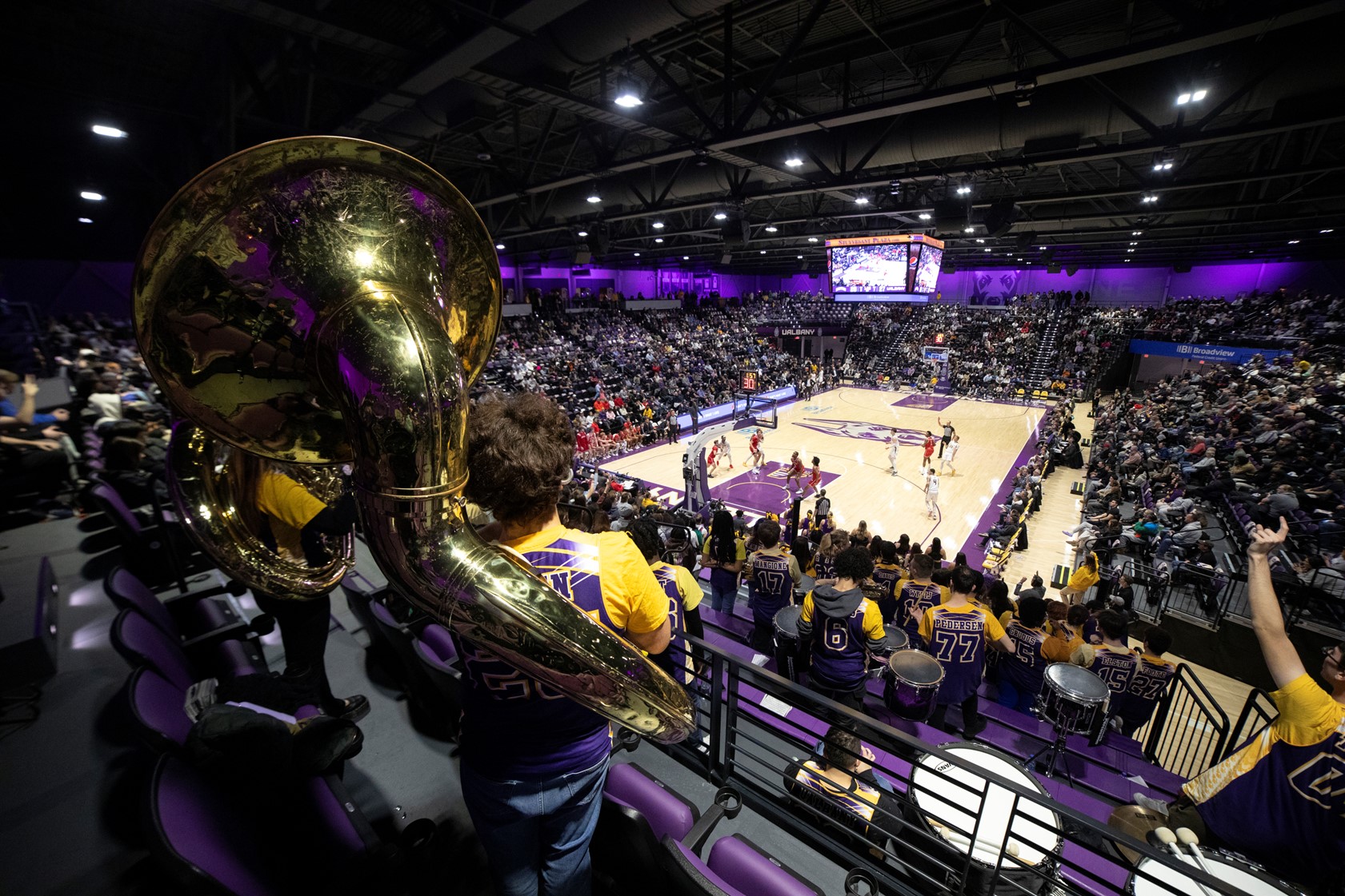A crowd cheers at a basketball game, the court lit up and the UAlbany band playing music.