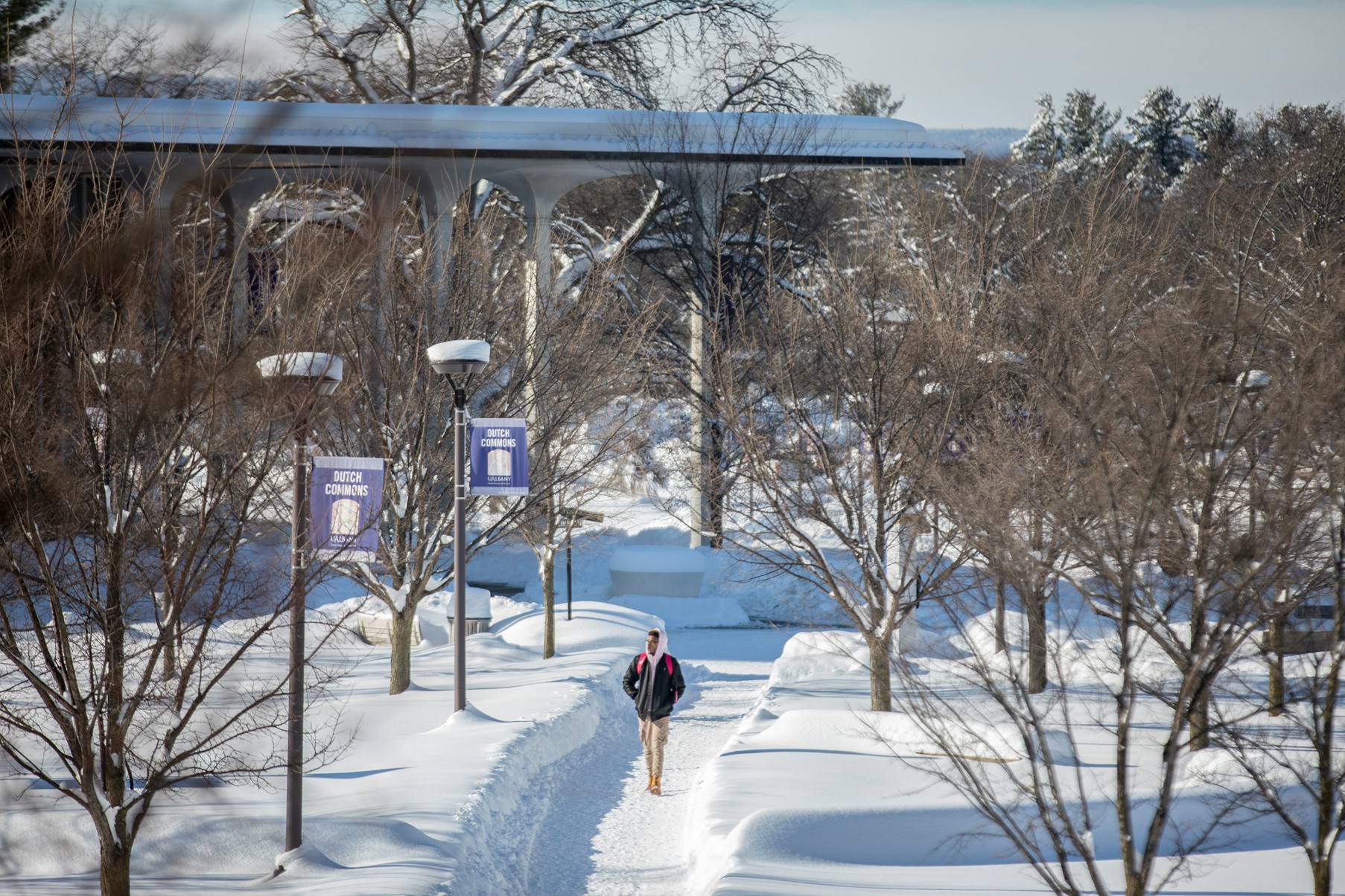 A person walks alone on campus in the winter. The snow has piled up high on the ground and they are surrounded by trees.
