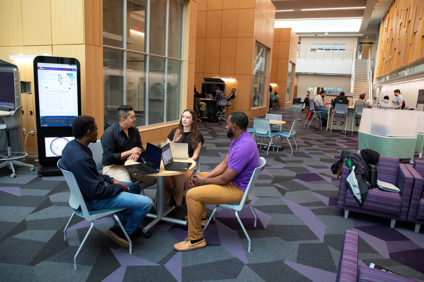 Four students sit around a round table in a Ualbany common space, talking to each other. There are other people in the background talking amongst each other.