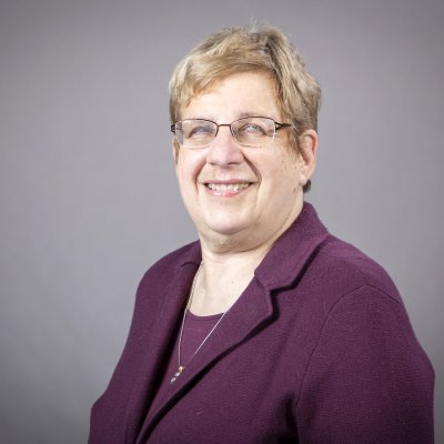 A woman with short blonde hair wearing glasses and a purple blouse and suit smiles for a portrait against a gray backdrop 