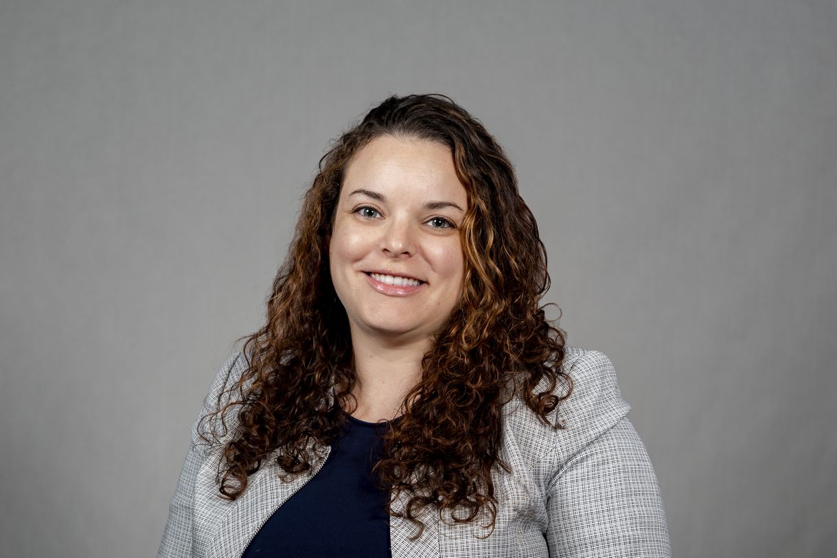 A woman with curly brown hair wearing a gray blazer and navy shirt smiles for a portrait in front of a gray backdrop.