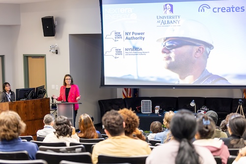 A woman in a hot pink blazer speaks in front of an auditorium of students in front of a large screen with a man wearing sunglasses and a hard hat.