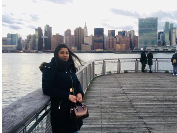 Farial Nur Maysha stands in a black coat on a wooden pier in front of a river and city skyline.